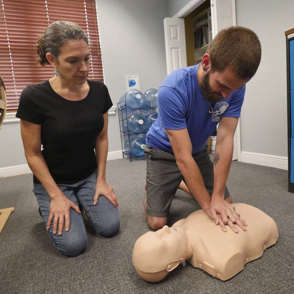 An employee practices workplace cpr aed while the patient instructor sits by them.