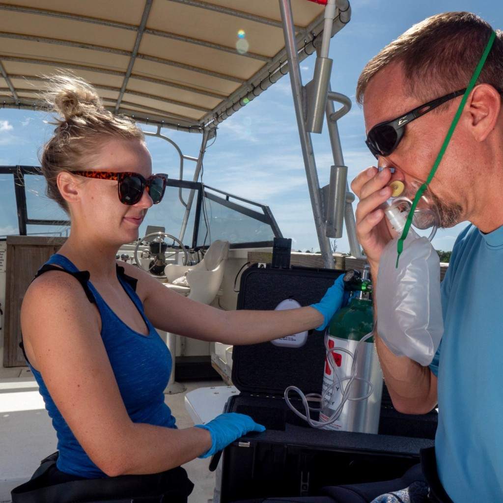 An individual trained in oxygen administration safely provides emergency O2 to a potentially injured diver through a non-rebreather mask on a boat.