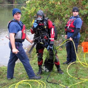 A public safety diver dressed in full face mask and bio-hazard dry suit with two PSD tenders in support.