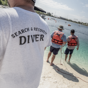Two divers on the shore acting as tenders, while another person in the foreground sports a Search & Recovery Diver tee shirt.